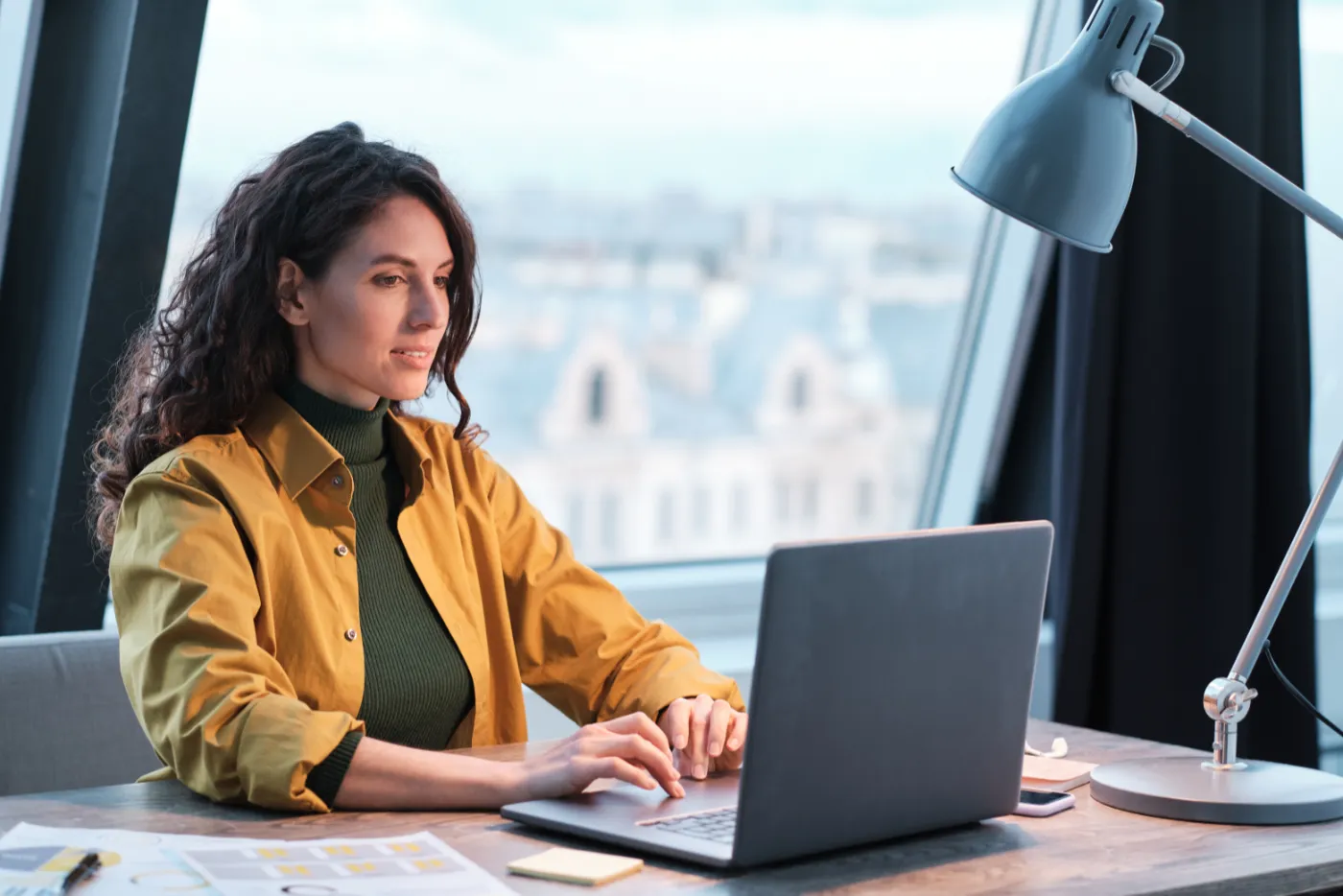 Woman typing on a laptop at a desk with city views in the background, focused on her work.