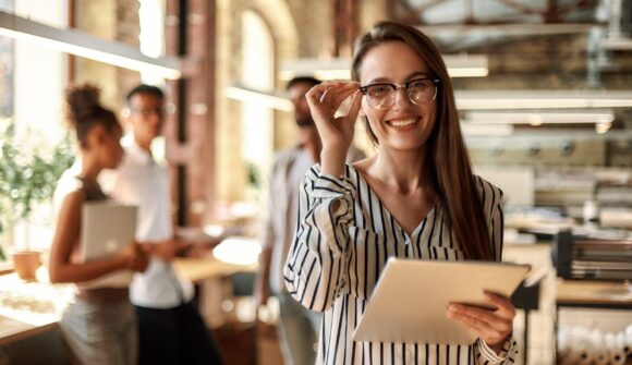 Successful business woman. Young cheerful woman holding digital tablet and looking at camera with smile while standing in the modern office. Business concept. Job
